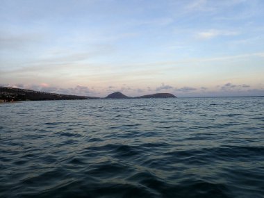 Alacakaranlıkta denizin yumuşak dalgalarını yakalayan sakin bir görüntü, Oahu, Hawaii 'deki Kahala Sahili' nde akşam gökyüzünün yumuşak renkleri altında uzak bir kıyı şeridi ve dağlar..