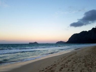 Waimanalo Sahili 'nde sakin ve güzel bir şafak manzarası. Nazik dalgalar el değmemiş kumları öper günün ilk ışıklarıyla gökyüzündeki yumuşak renklerin büyüleyici bir karışımını çizerken sakin suları ve uzak dağları aydınlatır.