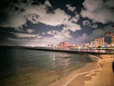 Gece Waikiki 'nin büyüleyici manzarası, yüksek binaların sakin plajla buluştuğu, ve okyanusun ritmik dalgalarının kentsel gelişim ve doğal güzelliğin harmanlanmış bir karışımını oluşturduğu yer. Işıldayan sular, canlı şehir ışıklarını yansıtıyor., 