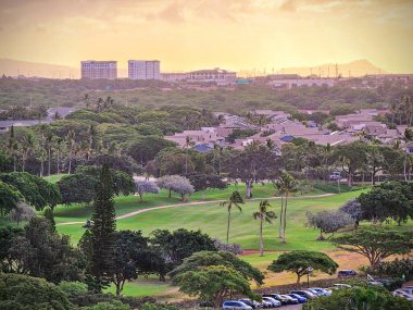 Oahu - 12 Mart 2024: Güneş ufkun altına batarken, Ko Olinas 'ın yemyeşil çayırları sıcak renklerle canlanıyor. Golf sahasında sakin bir akşam, uzakta Honolulus şehir ışıkları parıldıyor..  