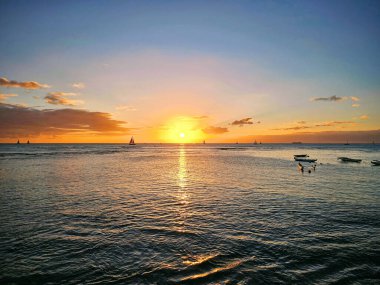 Güneş okyanusun üzerinde batıyor, suyun üzerine sıcak bir parıltı saçıyor. Ufuk boyunca yüzen birkaç tekne görülebilir, siluetleri Oahu, Hawaii 'deki renkli gökyüzüne karşıdır..