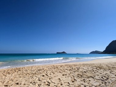 Waimanalo Sahili açıklarında berrak bir okyanusun yumuşak mavi dalgalarını öngören altın kumlarla dolu sakin bir manzara gözler önüne seriliyor. Ufuk küçük bir ada ile süslenirken, engin mavi gökyüzü neredeyse bulutsuzdur. Solda, kayalıklar usulca kumsalı kucaklıyor.