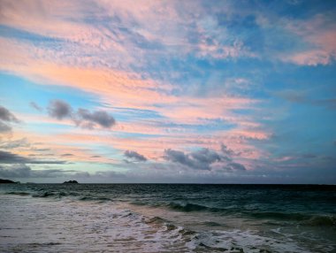 Güneş batarken gökyüzü yumuşak pastel renklerle parlıyor. Kıyıya vuran yumuşak okyanus dalgalarına güzel bir yansıma yansıtıyor..