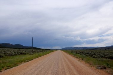 Sakin çakıllı bir yol, alçak tepelerle çevrili açık arazilere çıkar. Başında kara bulutlar belirir, muhtemel yağmuru işaret eder..