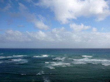 Dalgalar Oahu, Hawaii 'nin sahil şeridinde yavaşça çarpışıyor. Güneş öğleden sonra gökyüzünde dağınık bulutların arasından parlarken sakin bir atmosfer yaratıyor..