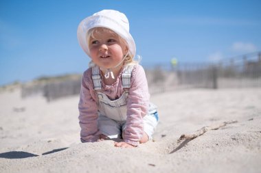 Küçük kız kumsalda oturuyor ve güneşli yaz gününde kumsalda oynuyor.