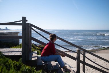 Middle-aged woman sitting on a wall looking at the ocean in Portugal, enjoying a sunny summer day. High quality photo