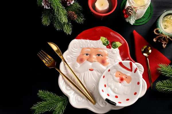 サンタクロースの頭の形のプレートと黒の表面にカトラリー キャンドルやモミの枝とボウルとクリスマステーブルの設定 — ストック写真