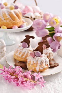 Easter muffins with icing sugar and bunny shaped cookies, ring cake and flowers on festive table in pastel colors 