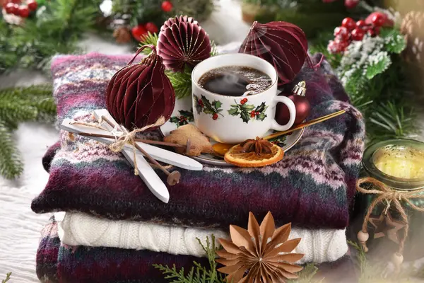 Winter coffee on stack of colorful woolen sweaters with burgundy paper ornaments and miniature ski decor