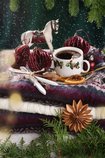 Winter coffee on stack of colorful woolen sweaters with burgundy paper ornaments and miniature ski decor with falling snow effect