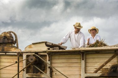 Pleudihen-sur-Rance, France, August 12, 2007: Two French farmers using ancient machine, threshing grain by removing seeds from the stalks and husks of hay on ancient combine harvester clipart