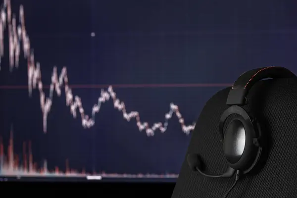 stock image black headset on chair over monitor with trading chart in background - dark background with selective focus and blur