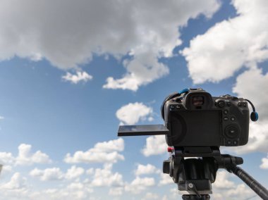 Yazın beyaz bulutlu mavi gökyüzünü gösteren bir tripodla çevirmeli ekranlı siyah profesyonel dijital kamera.