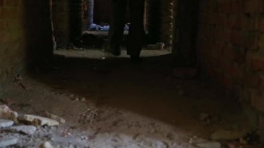 Yeşil asker pantolonlu biri, terk edilmiş karanlık binanın koridorunda dağınık tuğlalarla yürüyor.