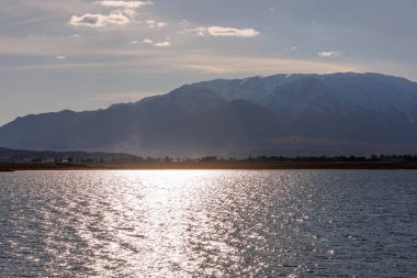 Issyk-Kul gölündeki mavi sakin su, güneşli bir sonbahar akşamında arka planda dağlar..