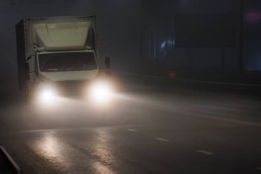 Otomotiv ışıklandırması olan bir araç gece yarısı sisli otoyolda ilerliyor. Fenerler asfalt yol yüzeyini aydınlatır ve karanlık gecede ilerler.