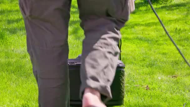 身穿塑料拖鞋的白人在阳光灿烂的日子用便宜的塑料电动除草机移动绿色草坪 用固定相机近距离观看 — 图库视频影像