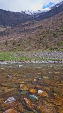 Geniş ama saydam bir dağ nehri. Altında çok sayıda yuvarlak kaya var. Arka planda karla kaplı dağ zirvesi var.