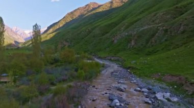 Bahar akşamları toprak yolu ve küçük deresi olan dağ geçidinin yeşil yamaçları alçak irtifa drone görüşü ile uçan hava aracı..