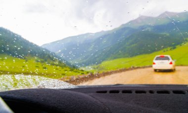 white car driving down the dirt road at rainy summer day in mountains, selective focus view from drivers perspective. clipart