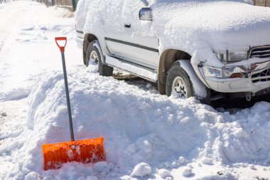 An orange snow shovel sticks out of a snowdrift in front of a snow-covered white SUV car on a sunny winter day clipart