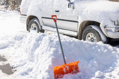 Turuncu bir kar küreği güneşli bir kış gününde kar kaplı beyaz bir SUV 'nin önünde kar yığınından sarkar.