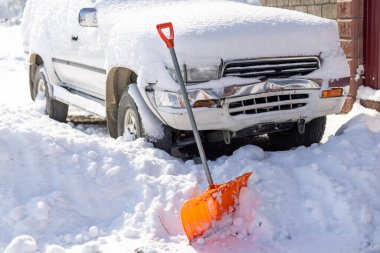 An orange snow shovel sticks out of a snowdrift in front of a snow-covered white SUV car on a sunny winter day clipart