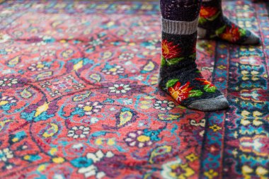Sıcak örülmüş yün çoraplar içinde kırmızı süslemeli halı zemininde pencere ışığıyla arka planda duran bacaklar.