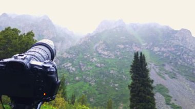 Siyah profesyonel bir kamera yağmurda bir dağ manzarası çekiyor, kameraya yağmur damlaları yağıyor - seçici bir odak ile yakın çekim