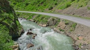 Kırgızistan 'da güneşli bir yaz gününde, dağ nehri boyunca uçarak dar dağ vadisinde boş toprak yolu ve yeşil yamaçlarla dolaşarak.