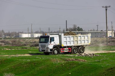 Beyaz Çin çöp kamyonu çamur ve yuvarlak kayalarla dolu güneşli yaz gününde endüstriyel bölgede toprak yol boyunca araba sürüyor.