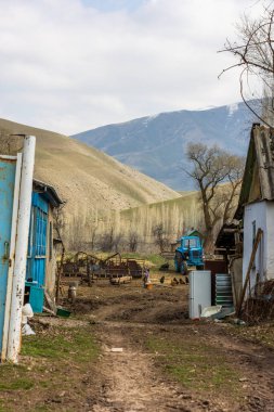 Güneşli bir bahar gününde paslı çelik kapının arkasındaki evlerin arasına toprak yolu olan eski mavi traktörüyle zavallı bir kır evi.