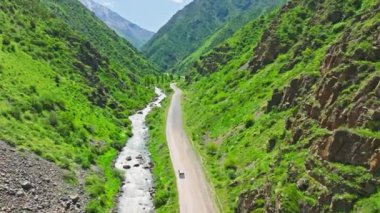 Gümüş minivan, Kırgızistan 'da güneşli bir yaz gününde, dar dağ vadisinde, dağ nehri kıyısındaki toprak yolda ilerliyor.