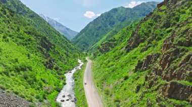 Gümüş minivan, Kırgızistan 'da güneşli bir yaz gününde, dar dağ vadisinde, dağ nehri kıyısındaki toprak yolda ilerliyor.