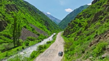 Kırgızistan 'ın güneşli yaz gününde yeşil yamaçlı dar dağ vadisinde, dağ nehri kıyısındaki toprak yolda araba ilerliyor.