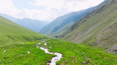 Kırgızistan 'da güneşli bir günde çim yamaçlı yeşil dağ geçidi ve örülmüş nehir. Tian-Shan dağ sırasının üzerinde beyaz bulutlu mavi gökyüzü, Dolly ileri kamera hareketli drone bakış açısı.