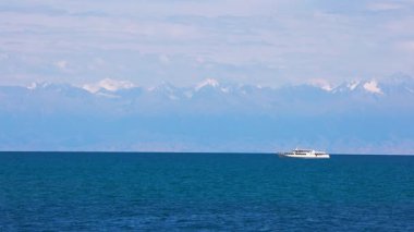 Kırgızistan 'daki Issyk-Kul Gölü' nde beyaz bir yolcu teknesinin arka planında büyüleyici karla kaplı dağlar var. Sakin mavi su ve berrak gökyüzü huzurlu ve manzaralı bir manzara yaratır..
