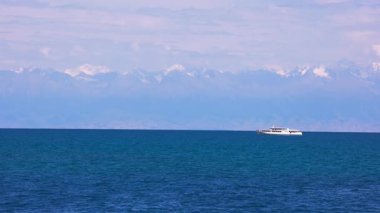 Kırgızistan 'daki Issyk-Kul Gölü' nde beyaz bir yolcu teknesinin arka planında büyüleyici karla kaplı dağlar var. Sakin mavi su ve berrak gökyüzü huzurlu ve manzaralı bir manzara yaratır..