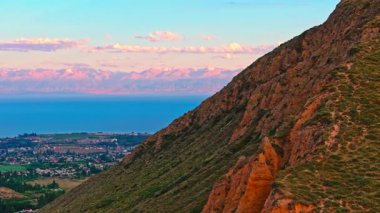 Dağ gölünün kıyısında ufukta karla kaplı bir dağ silsilesi olan küçük bir kasaba yaz günbatımında drone atışını gözler önüne seriyor.