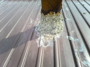 Kahverengi metal çatıdaki paslanmış çelik bir soba borusunun yakın plan görüntülerinde çatlaklar, pas ve zayıf mühür görülüyor. Çatı tamiri, boru takviyesi ve sızıntıları önlemek için uygun mühür ihtiyacını gösterir..