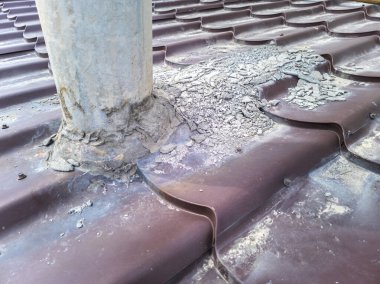 Kahverengi metal çatıdaki paslanmış çelik bir soba borusunun yakın plan görüntülerinde çatlaklar, pas ve zayıf mühür görülüyor. Çatı tamiri, boru takviyesi ve sızıntıları önlemek için uygun mühür ihtiyacını gösterir..