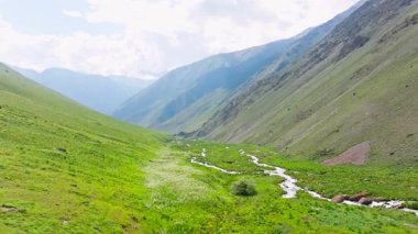 Kırgızistan 'da güneşli bir günde çim yamaçlı yeşil dağ geçidi ve örülmüş nehir. Tian-Shan dağ sırasının üzerinde beyaz bulutlu mavi gökyüzü, Dolly ileri kamera hareketli drone bakış açısı.
