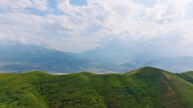Güneşli bahar gününde, dağların önündeki çimenli yeşil tepelerin hava manzarası, parallax efektli kamyon kamerası hareketi.