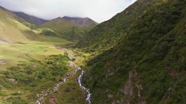 Bir dron, yemyeşil tepelerin arasından akan berrak bir nehri olan manzaralı bir dağ vadisini yakalar. Manzara kayalık arazi, yoğun bitki örtüsü ve bulutlu bir gökyüzünün altında sisli zirveler içerir..