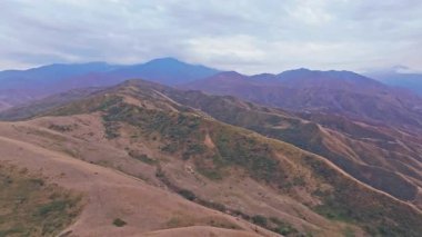 Kırgızistan 'daki Chu Vadisi' nin engebeli eteklerini görüntüleyen hava görüntüleri, kayan tepeleri, katmanlı dağları ve bulutlu gökyüzünü gösteriyor, kamera sürekli olarak doğal manzara üzerinde hareket ediyor..