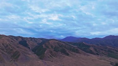 Hava görüntüleri engebeli bir dağ manzarasını derin vadilerle ve parçalı bulutlu gökyüzüyle yakalıyor. Kamera aşağıya doğru hareket ediyor, engin, kurak araziyi ve sakin, doğal ortamdaki uzak tepeleri gösteriyor..