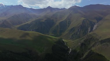 İnsansız hava aracı uzak bir Kırgızistan vadisindeki Tuyuk Nehri üzerinde uçuyor. Karla kaplı tepeleri ve bulutlu gökyüzünün altındaki yeşil tepeleri olan engebeli dağlar..