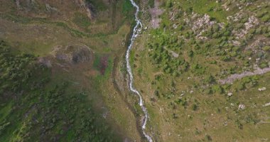 İnsansız hava aracı, dağılmış ağaçlar ve kayalık arazisi olan yeşil bir vadiden geçen bir dereyi yakalıyor. Kamera sola döner, dikey olarak aşağı bakar ve doğal manzarayı gösterir..