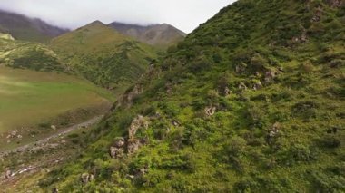 Bir dron, yemyeşil tepelerin arasından akan berrak bir nehri olan manzaralı bir dağ vadisini yakalar. Manzara kayalık arazi, yoğun bitki örtüsü ve bulutlu bir gökyüzünün altında sisli zirveler içerir..